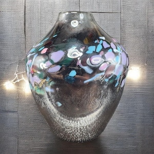 May include: A decorative glass vase with a silver metallic finish. The vase features colorful, abstract accents in shades of blue, pink, and purple. The vase has a textured base and a wide, rounded shape.