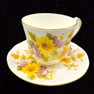 May include: A white teacup and saucer with a floral pattern. The pattern features yellow and purple flowers on a white background with a gold trim.