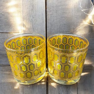 May include: Two clear glass tumblers with a yellow honeycomb pattern. The glasses have a rounded shape and are illuminated by sunlight, creating a warm glow. The honeycomb design is a repeating geometric pattern, adding a decorative element to the glassware.