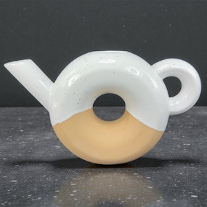 Peut inclure: Une th&eacute;i&egrave;re en c&eacute;ramique en forme de donut. La moiti&eacute; sup&eacute;rieure est blanche avec des d&eacute;tails mouchet&eacute;s, tandis que la moiti&eacute; inf&eacute;rieure est de couleur beige clair. Elle comporte un bec verseur et une poign&eacute;e arrondie.