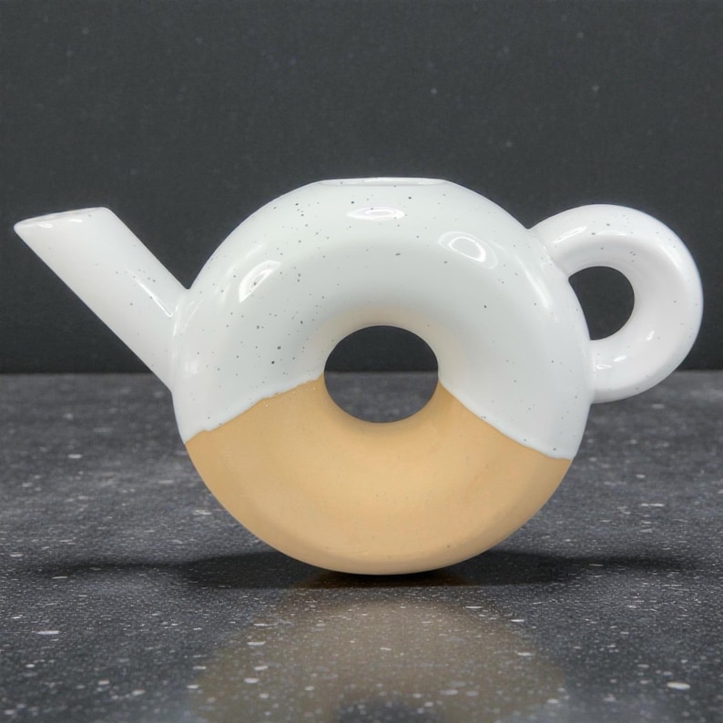 Peut inclure: Une th&eacute;i&egrave;re en c&eacute;ramique en forme de donut. La moiti&eacute; sup&eacute;rieure est blanche avec des d&eacute;tails mouchet&eacute;s, tandis que la moiti&eacute; inf&eacute;rieure est de couleur beige clair. Elle comporte un bec verseur et une poign&eacute;e arrondie.