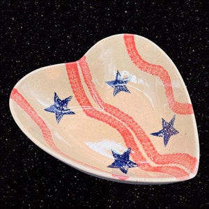 May include: A heart-shaped ceramic bowl with a cream-colored base and red stripes. The bowl is decorated with four blue stars.