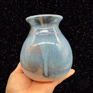 May include: A small, round, blue and white ceramic vase with a glossy finish. The vase has a subtle, textured pattern.