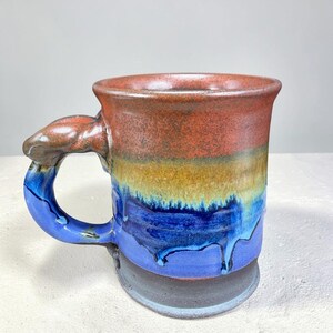 May include: Handmade ceramic mug with a unique handle design. The mug features a gradient of colors, including red, yellow, and blue. The handle is brown, and the base is a darker shade of blue. The mug is a functional piece of art.