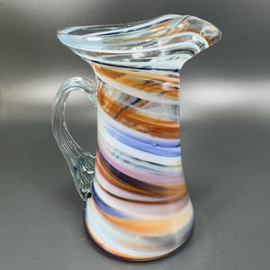 May include: A hand-blown glass pitcher with a clear handle and a flared top. The pitcher features a colorful swirl design of orange, white, blue, and pink. The pitcher is approximately 8 inches tall.
