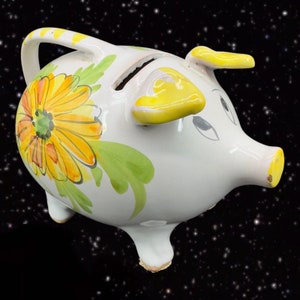 May include: White ceramic piggy bank with a yellow ear and a floral design featuring yellow and orange flowers. The piggy bank has a slot on the top for coins.