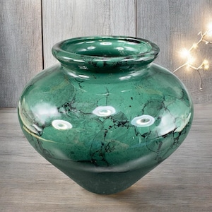 May include: A green decorative vase with a rounded body and a wide opening. The vase has a glossy finish with black veining throughout. The vase is made of a solid material and sits on a wooden surface.
