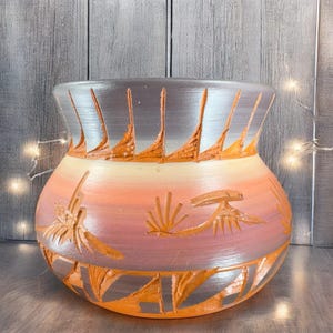 May include: A large, orange, brown, and silver ceramic pot with a wide mouth and a rounded body. The pot has a carved design of stylized birds and plants.