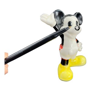 K&ouml;nnte beinhalten: Eine wei&szlig;e Keramikfigur von Mickey Mouse mit schwarzen Ohren, schwarzen Schuhen und einem rot-schwarzen Outfit. Mickey winkt mit der rechten Hand und ein schwarzer Stift zeigt auf ihn.