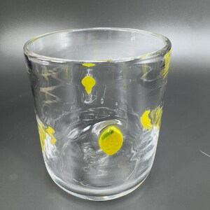 May include: A clear glass tumbler with yellow lemon designs. The glass has a slightly textured surface and a wide, cylindrical shape. The yellow lemon designs are scattered around the glass, adding a playful touch to the glassware.