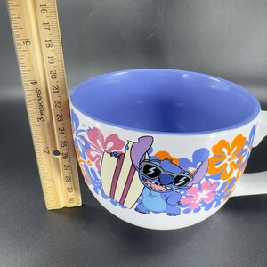 May include: A white ceramic mug with a blue and orange floral design. The mug features a cartoon character of Stitch from Lilo & Stitch wearing sunglasses and holding a surfboard. The inside of the mug is blue.