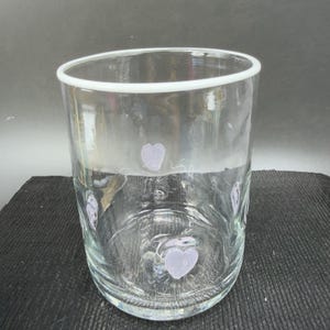 May include: A clear glass tumbler with a white rim and lavender heart-shaped designs. The glass is cylindrical and appears to be empty. The hearts are evenly spaced around the glass.