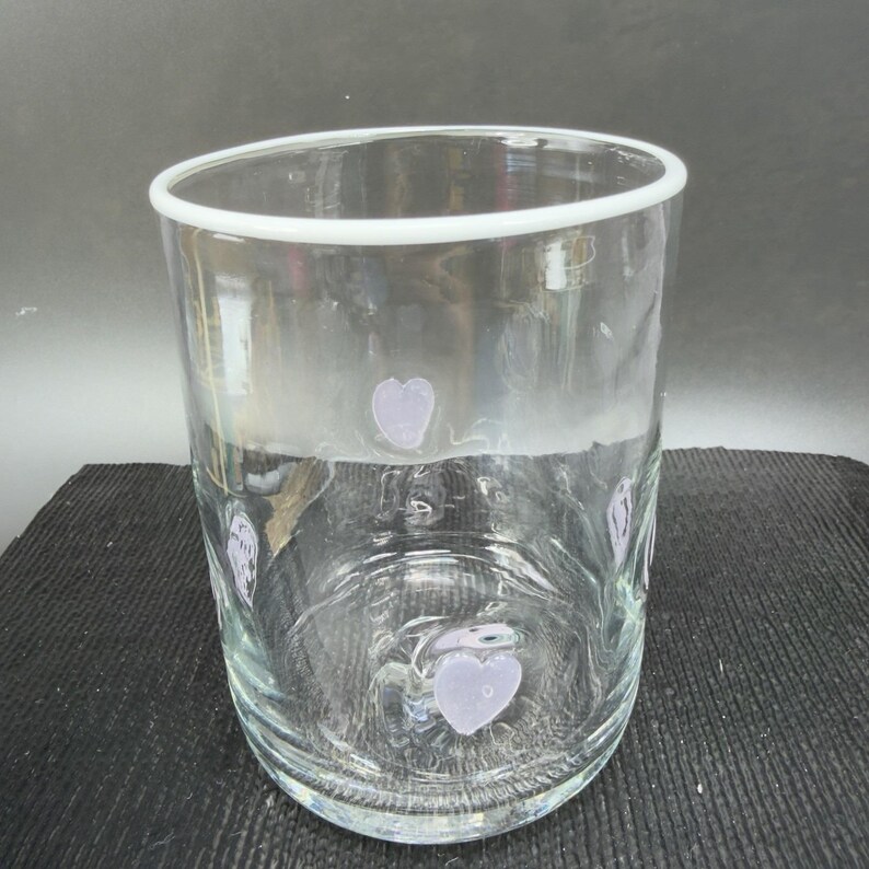 May include: A clear glass tumbler with a white rim and lavender heart-shaped designs. The glass is cylindrical and appears to be empty. The hearts are evenly spaced around the glass.