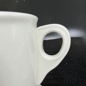 May include: A white ceramic mug with a black handle and the text "But First, Coffee" in a handwritten style. The mug is on a white surface. The mug is approximately 4 inches tall.