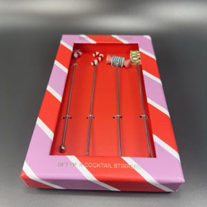 May include: A set of four cocktail stirrers with candy-themed toppers, presented in a red and pink striped box. The stirrers have various designs, including a swirl, a candy cane, and a wrapped candy. The box reads "SET OF 4 COCKTAIL STIRRERS".