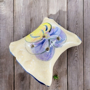 May include: A ceramic soap dish shaped like a pillow with a hand-painted purple and yellow flower design. The dish has several drainage holes.