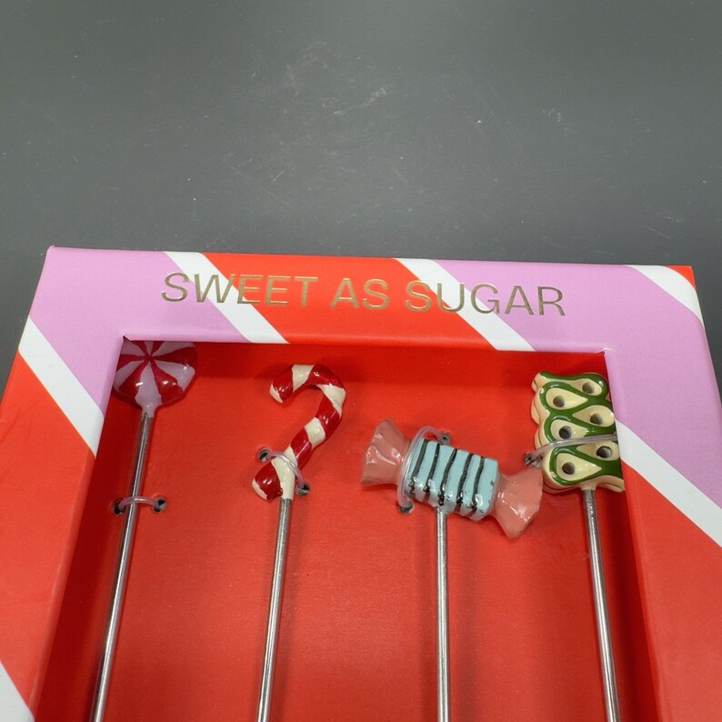 May include: A set of four decorative candy-themed skewers. The skewers feature a lollipop, candy cane, wrapped candy, and a green and yellow design. The box is red and pink with the text "SWEET AS SUGAR".