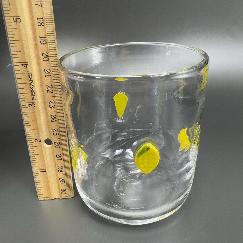 May include: Clear glass tumbler with yellow lemon-shaped designs. The glass is cylindrical with a slightly flared top. A ruler is placed next to the glass, showing measurements in inches.