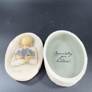 May include: White ceramic trinket box with an angel holding a bouquet of purple flowers. The lid of the box has the words "Thank You" inscribed on it. The inside of the box has the words "Appreciating your kindness!" written in black ink.
