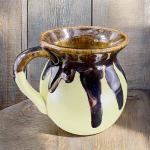 May include: A yellow ceramic mug with a brown and black glaze. The mug has a rounded body and a handle.