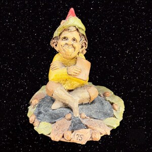May include: A small ceramic figurine of a gnome wearing a red and green hat, sitting on a leaf-shaped base. The gnome is holding a book and has a friendly expression.