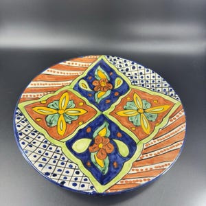 May include: A round ceramic plate with a colorful floral design. The plate features four diamond-shaped sections with blue, green, yellow, and orange flowers on a white background. The plate has a white border with a black and white polka dot pattern.
