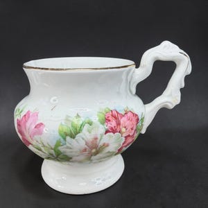 May include: White ceramic teacup with a floral design featuring pink and white flowers and green leaves. The cup has a gold rim and a decorative handle. The teacup sits on a small pedestal base.