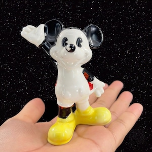 K&ouml;nnte beinhalten: Eine wei&szlig;e Keramikfigur von Mickey Mouse mit schwarzen Ohren und Schuhen, roten Shorts und einem gelben L&auml;cheln. Die Figur winkt mit dem rechten Arm.