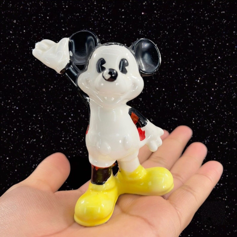 K&ouml;nnte beinhalten: Eine wei&szlig;e Keramikfigur von Mickey Mouse mit schwarzen Ohren und Schuhen, roten Shorts und einem gelben L&auml;cheln. Die Figur winkt mit dem rechten Arm.