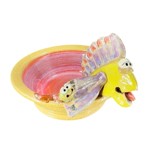 May include: A whimsical ceramic bowl in the shape of a fish. The bowl is yellow with a red and pink interior. The fish has a yellow body, purple fins, and a red tongue. The bowl is a unique decorative piece.