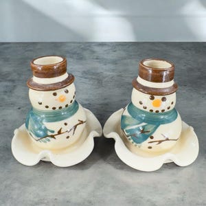May include: Two ceramic snowman candle holders with brown hats, blue scarves, and white and brown accents. The candle holders are sitting on a white surface.