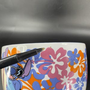 May include: White ceramic mug with a blue, pink, and orange floral pattern. Stitch, a blue alien character with sunglasses, is featured on the mug.