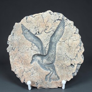 May include: A circular, textured, stone-like wall hanging featuring a detailed relief of a seagull in flight. The bird is gray, with outstretched wings, set against a light brown background with a cracked appearance. The artwork is mounted on a white stand.