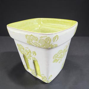 May include: A square ceramic planter with a white exterior featuring a light green floral pattern. The interior is glazed in a bright lime green. The base of the planter includes vertical drainage slots on each side of the square body.