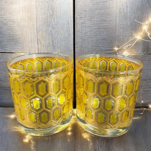 May include: Two clear glass votive candle holders with a yellow honeycomb pattern. The honeycomb design is outlined in gold. The candle holders are cylindrical and sit on a wooden surface.