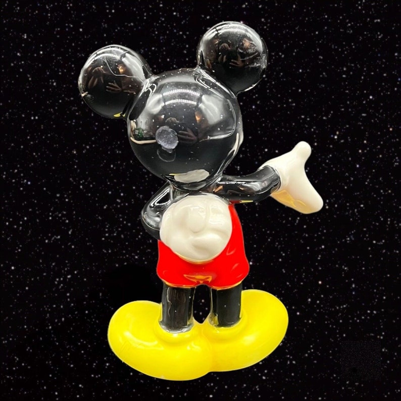 Puede incluir: Una figurita de cer&aacute;mica negra y blanca de Mickey Mouse con pantalones cortos rojos y zapatos amarillos. Est&aacute; de pie con el brazo derecho extendido y la mano izquierda en la cadera.