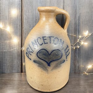 May include: Tan stoneware jug with a handle, featuring the text "PRINCETON, ILL." in blue, along with a blue heart design. The jug has a rustic, aged appearance, suggesting it is vintage or antique.