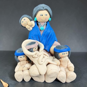 May include: A clay figurine depicting a central figure with gray hair wearing a blue shawl and turquoise jewelry, holding a small infant. Three additional small figures with dark hair and blue headbands are positioned around the base.