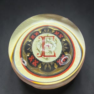 May include: A circular glass paperweight featuring a central cream-colored medallion with a large red letter E surrounded by intricate green vine illustrations. The medallion is framed by a black border with red floral accents and gold details.