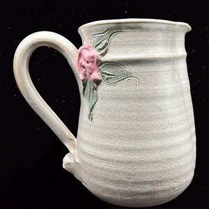 May include: A white ceramic pitcher with a floral design. The pitcher has a handle and a wide mouth. The floral design features a pink flower with green leaves.
