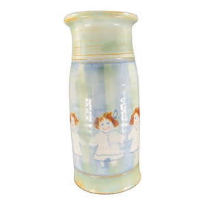 May include: A light blue ceramic vase with a striped pattern and hand-painted images of dolls. The dolls are wearing white dresses and have red hair.