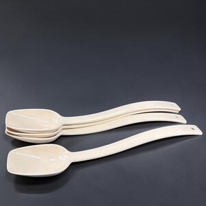 May include: A set of three cream-colored plastic spoons with square-shaped bowls and long, curved handles. Each handle features a small circular hole at the end, designed for hanging. The spoons are stacked neatly, showcasing their smooth finish.