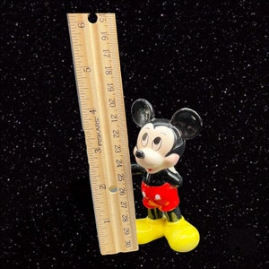 Puede incluir: Una figurita de cer&aacute;mica en blanco y negro de Mickey Mouse. Mickey lleva pantalones cortos rojos con un bot&oacute;n amarillo y zapatos amarillos. Est&aacute; de pie junto a una regla de madera.