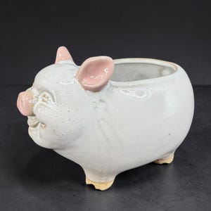 May include: A ceramic planter shaped like a pig. The planter is light gray with pink accents on the ears and snout. It has a round body and three small feet. The pig is smiling, and the planter is suitable for small plants or succulents.