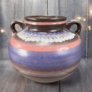 May include: A round, ceramic vase with two handles. The vase features horizontal bands of color, including brown, pink, purple, and white. The top rim is dark brown, and the design includes a pattern of white and blue dots.