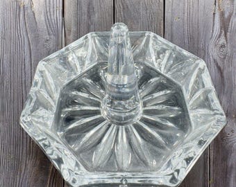 May include: A clear glass ring holder with a decorative, octagonal design and a raised center post. The holder has a textured, faceted surface.