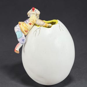 May include: A whimsical figurine of a clown emerging from a large white egg. The clown is dressed in a yellow and patchwork outfit with a red nose. The egg has a cracked design, and the interior is yellow.
