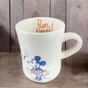 May include: White ceramic mug with a blue and red illustration of Mickey and Minnie Mouse. The mug has the text "fun times!" written in orange on the inside.