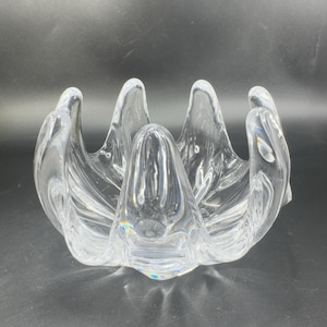 May include: Clear glass bowl with a unique, organic design. The bowl features a central, rounded form surrounded by petal-like extensions. The glass is transparent, allowing light to pass through, and the bowl sits on a dark surface.