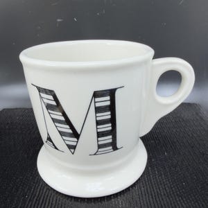 May include: White ceramic mug with a black, striped letter "M" on the front. The mug has a rounded base and a handle. The mug is on a dark surface.